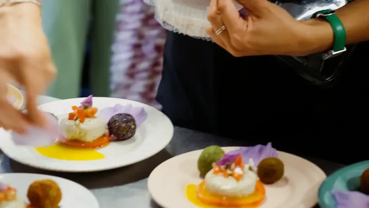 Cheffe en tablier noir dressant des assiettes de cuisine végétale à Marseille, une fleur comestible mauve à la main : dômes, billes colorées et sauces jaunes, école ORA © Geoffrey Roy