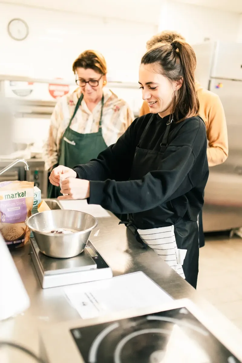 Participante d'un stage de cuisine végétale à Marseille pesant un ingrédient dans un saladier, sa cheffe en arrière-plan, école ORA © Julie Vandal