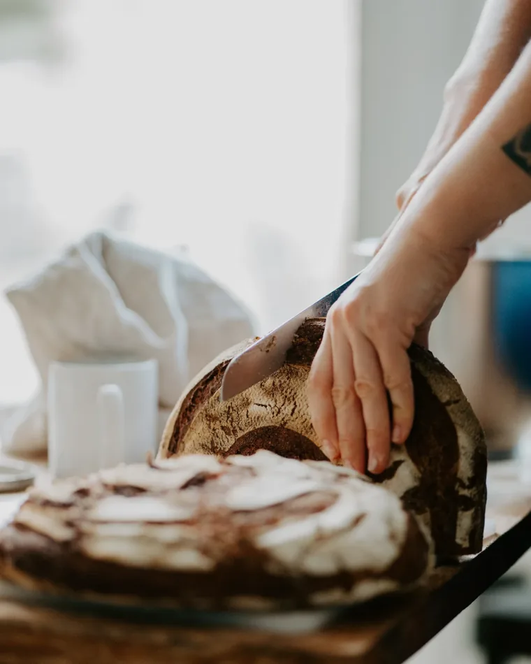 Mains tranchant au couteau une miche de pain au levain à la mie marbrée, posée sur une table en bois, école ORA © chefJM