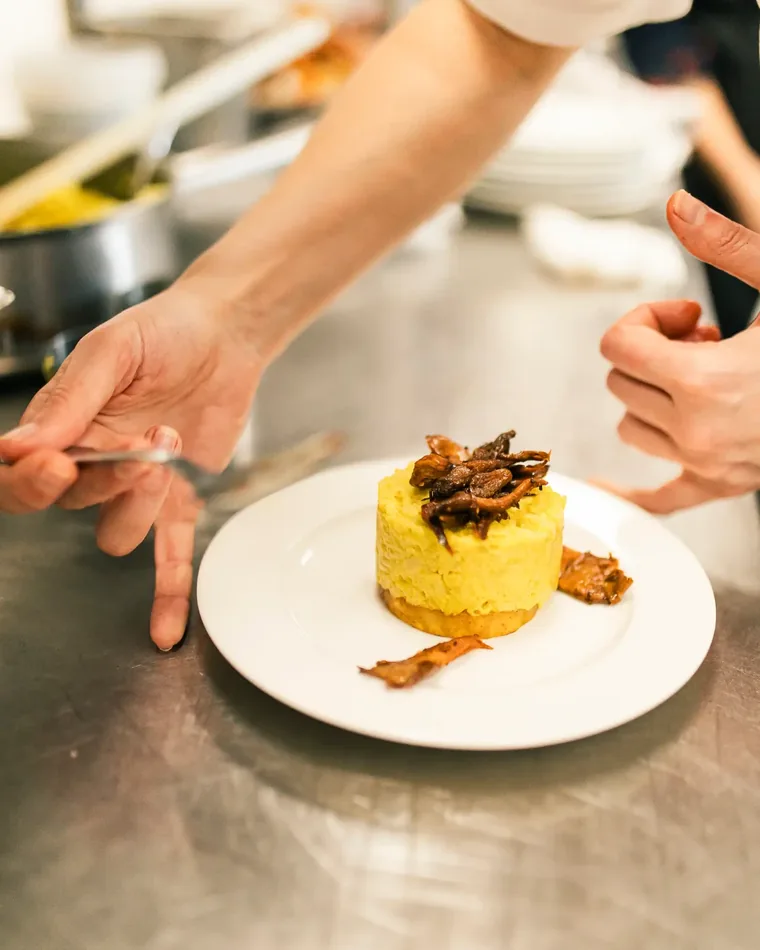 Cheffe en uniforme blanc dressant une assiette de cuisine végétale : dôme de purée de légumes surmonté de champignons sautés, école ORA © Julie Vandal