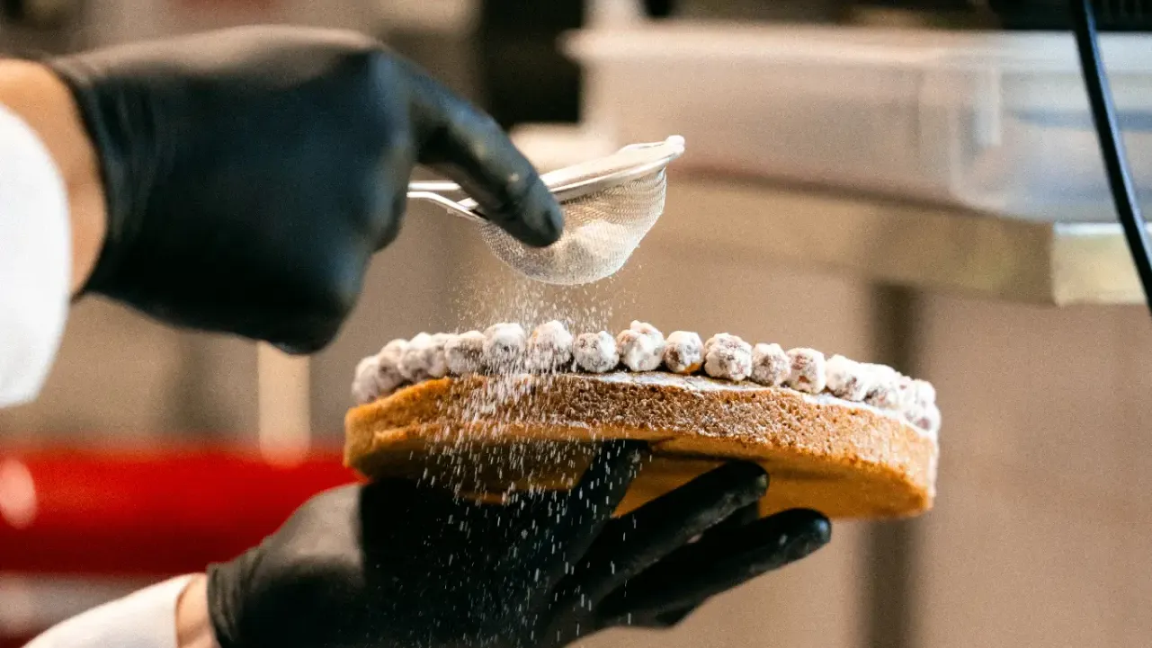 Mains gantées saupoudrant de sucre glace un dessert végétal à l'aide d'un tamis, en atelier de pâtisserie